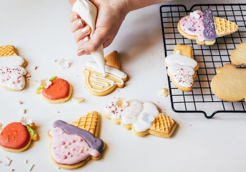 A hand decorates ice cream-shaped cookies with icing. More decorated cookies and a cooling rack are visible.