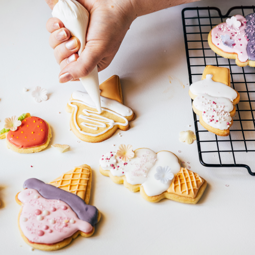 A hand decorates ice cream-shaped cookies with icing. More decorated cookies and a cooling rack are visible.