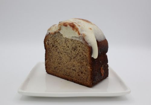 A slice of banana bread with icing on top, resting on a white plate against a plain background, showcasing a delicious homemade treat.