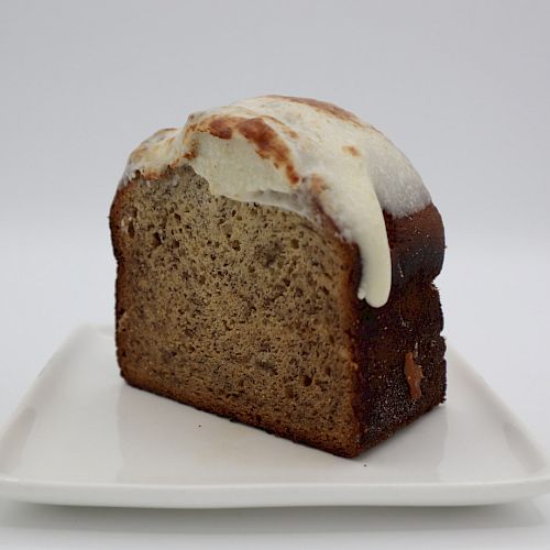 A slice of banana bread with icing on top, resting on a white plate against a plain background, showcasing a delicious homemade treat.