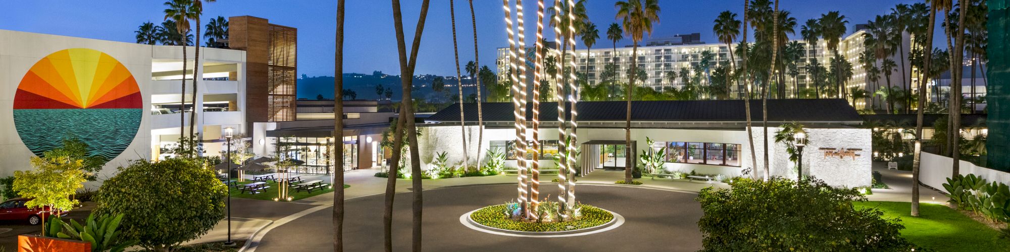 A landscaped entrance with palm trees wrapped in lights, vibrant signage, and a sunset mural on a white building.